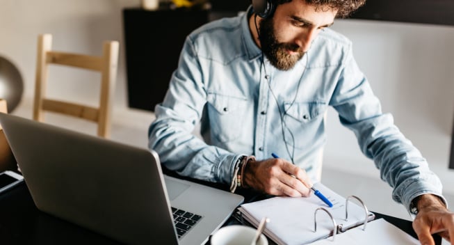 Portrait of busy bearded man in denim shirt and headphones checking planner at table with laptop.