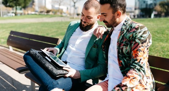 Portrait of bearded men in bright jackets browsing tablet on bench in sunny park.