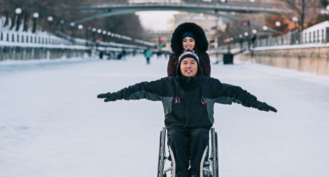 Man in wheelchair and woman skating on canal.