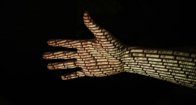 Hand In Front Of Black Background With Binary Code Projected Onto The Skin.