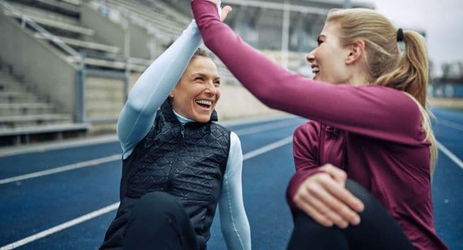 Two laughing women sitting on a track high fiving together after a run