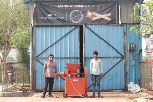 Two studnets stand in front of a blue metal warehouse and display a bright orange piece of machinery. 