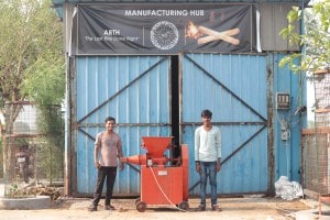 Two studnets stand in front of a blue metal warehouse and display a bright orange piece of machinery.