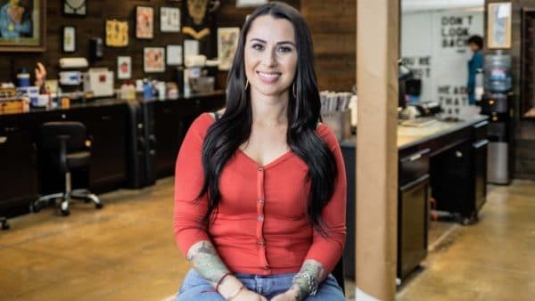 A smiling woman with long brown hair and arm tattoos wearing a red top and blue jeans sits in a workshop.