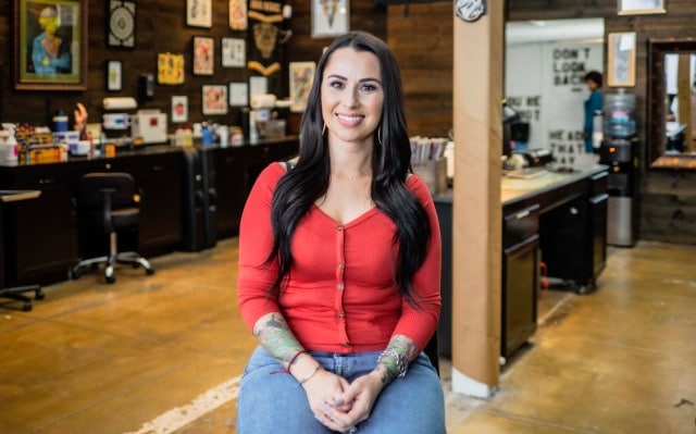 A smiling woman with long brown hair and arm tattoos wearing a red top and blue jeans sits in a workshop.