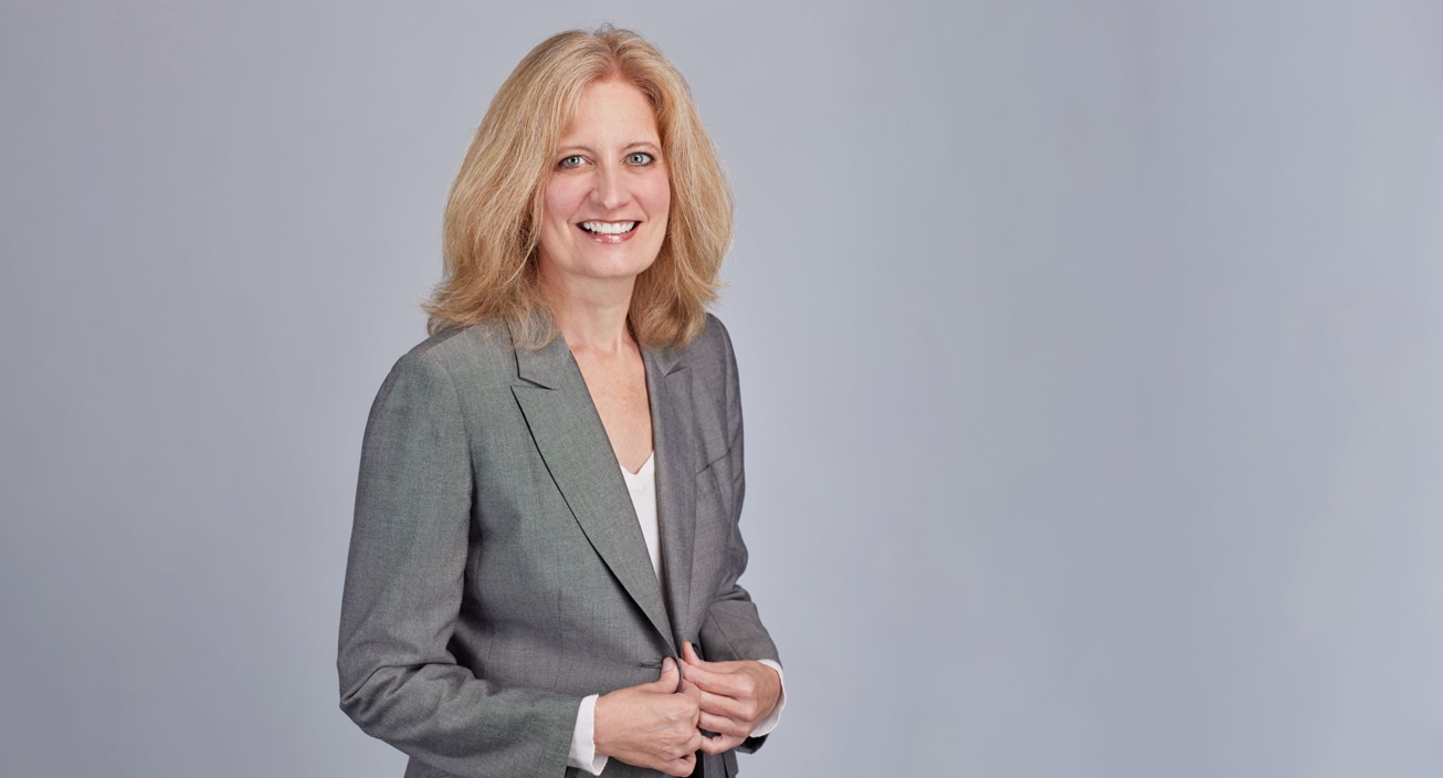 A blonde woman in a grey suit poses for a professional photo in front of a grey background.