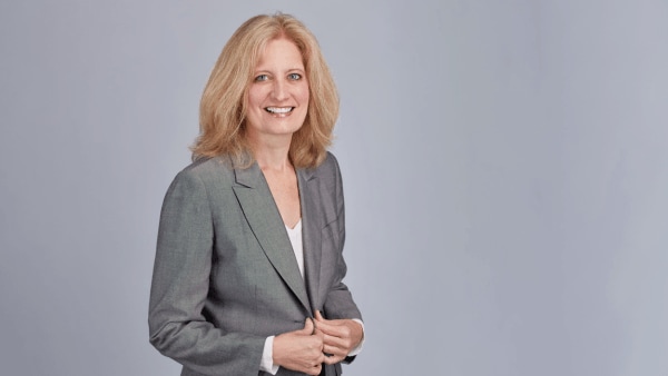 A blonde woman in a grey suit poses for a professional photo in front of a grey background.