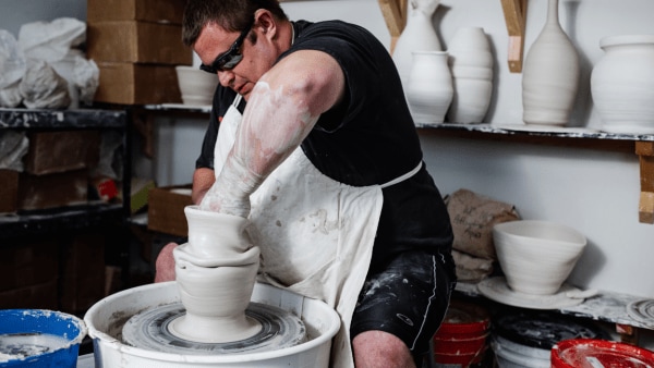 A man wearing sunglasses and a white apron molds clay on a potters wheel with finished vases and bowls in the background.