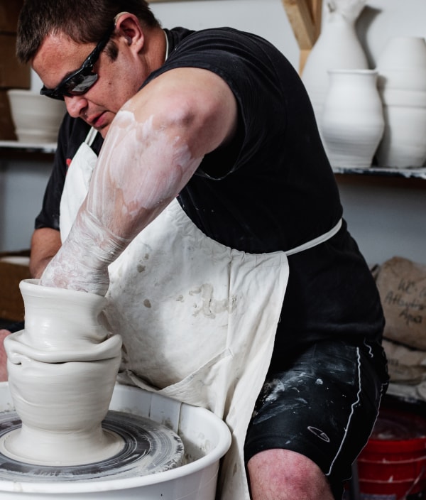 A man wearing sunglasses and a white apron molds clay on a potters wheel with finished vases and bowls in the background.