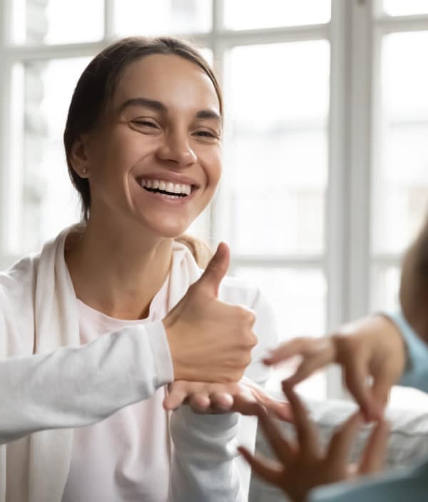 A smiling female wearing a white hoodie uses ASL to communicate.