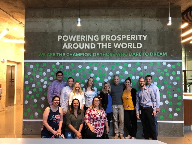 Intuit employees pose in front of a cement wall with white and green polka dots with the words, "Powering Prosperity Around the World," above the group. 