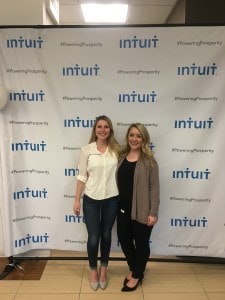 Two blonde women in professional clothes pose in front of a white banner featuring the Intuit logo.