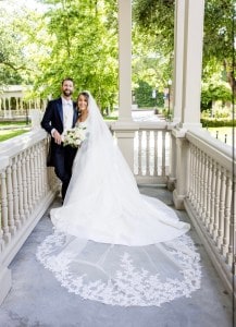 A bride wearing a long veil with delicate lace around the edge poses with her husband.