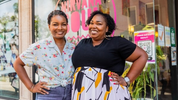 Two smiling Black women pose in front of their coffee shop with a pink window decal promoting Nirvana Soul.