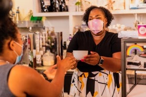 Black female coffee shop owner serves a customer while wearing a mask.