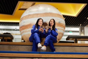 Two women in blue coveralls resembling astronauts pose in front of a fake Jupiter with heart-shaped churros.