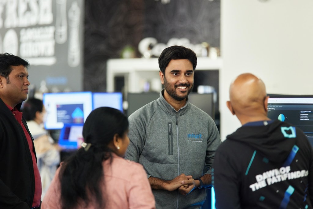 A technologist stands by his computer while smiling and hearing pitches from other team members. 
