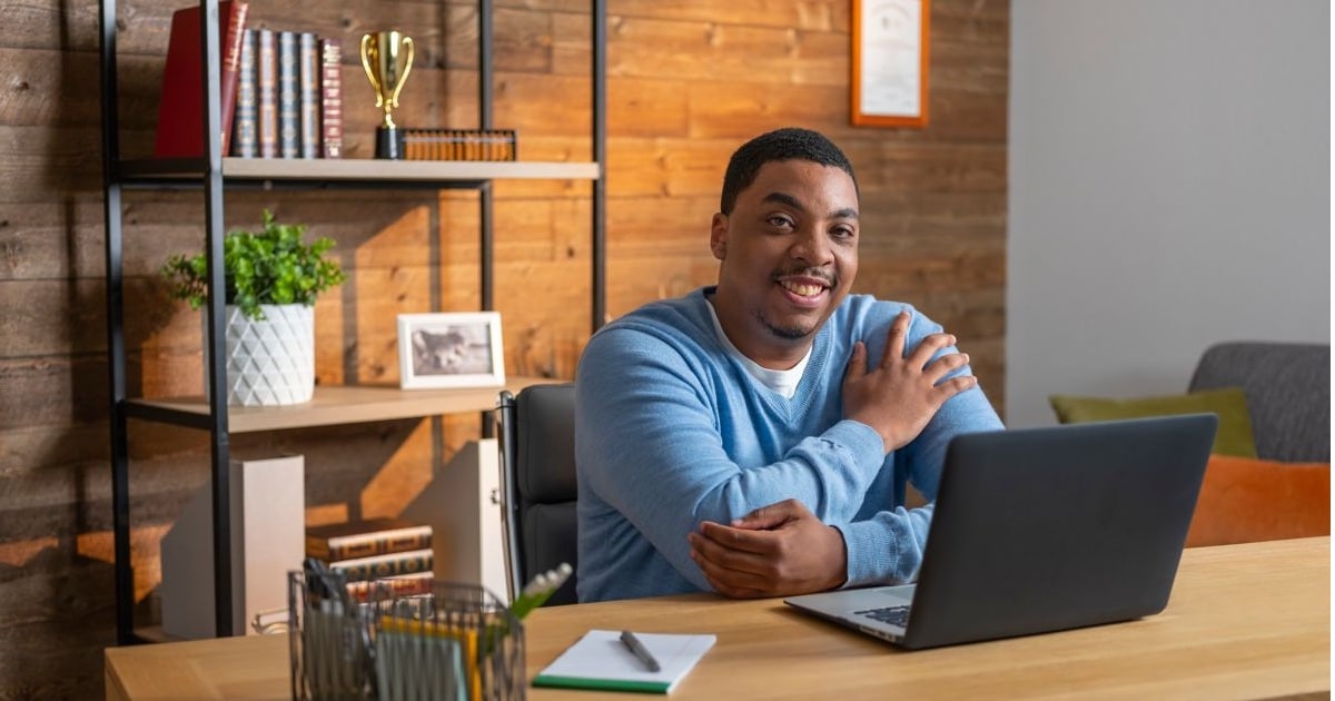 A man in a blue sweather smiles while working at his open laptop in a nicely adorned, masculine-looking office.