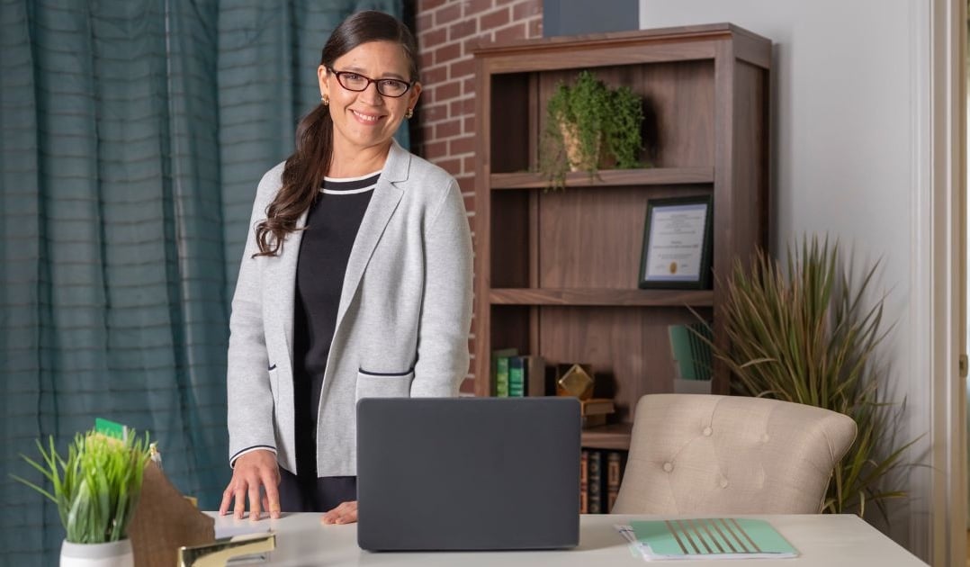 A business woman poses in her nicely furnished office wearing glasses and a blazer.