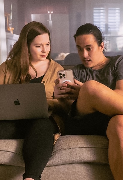 A woman in a yellow sweater sits on her couch with her computer open on her lap. Next to her a man with black hair in gym attire shows her something on his phone.