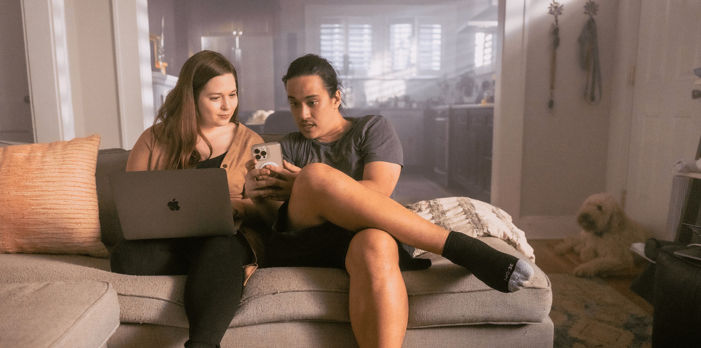 A woman and man sit on the couch with an open laptop while looking a a phone.