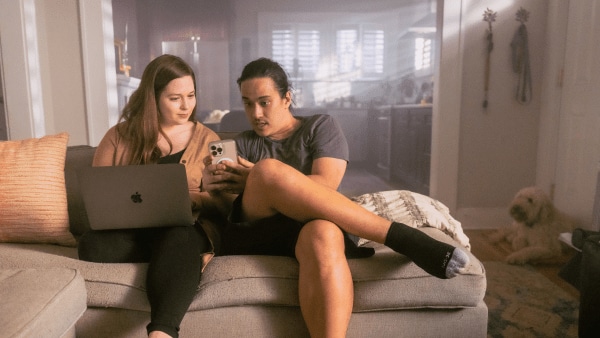 A woman and man sit on the couch with an open laptop while looking a a phone.