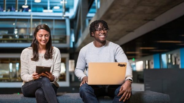 A brunette tech worker in her 20s smiles while looking at her tablet in an industrial chic open office setting. Next to her a young, Black man with short braids works at his computer.