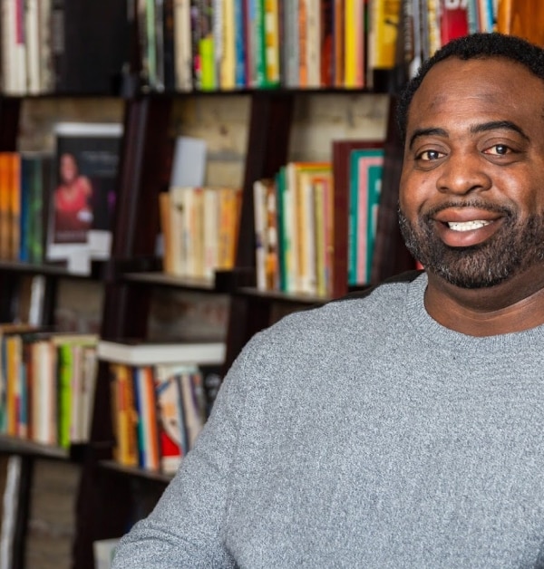 A professional headshot of a Black man in a grey sweater, smiling at the camera with a colorful bookcase in the background.