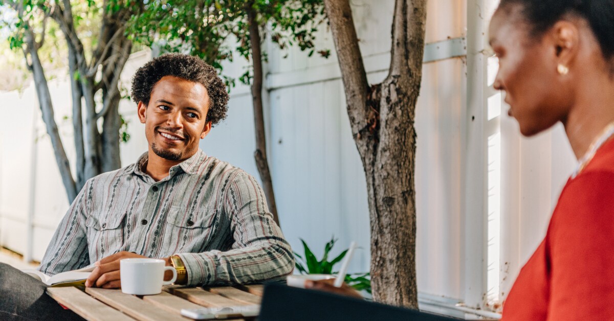 A black man in a button down shirt smiles at his wife while drinking coffee outside.