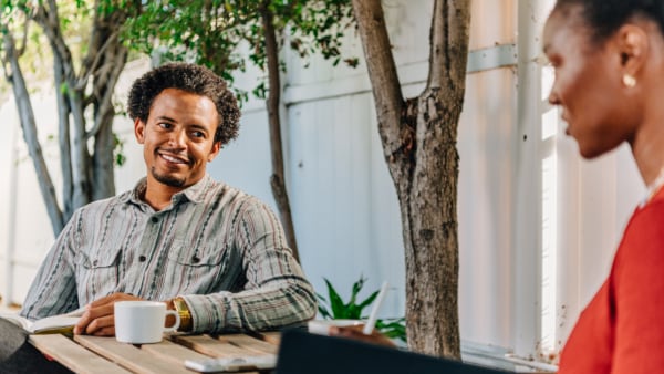 A black man in a button down shirt smiles at his wife while drinking coffee outside.