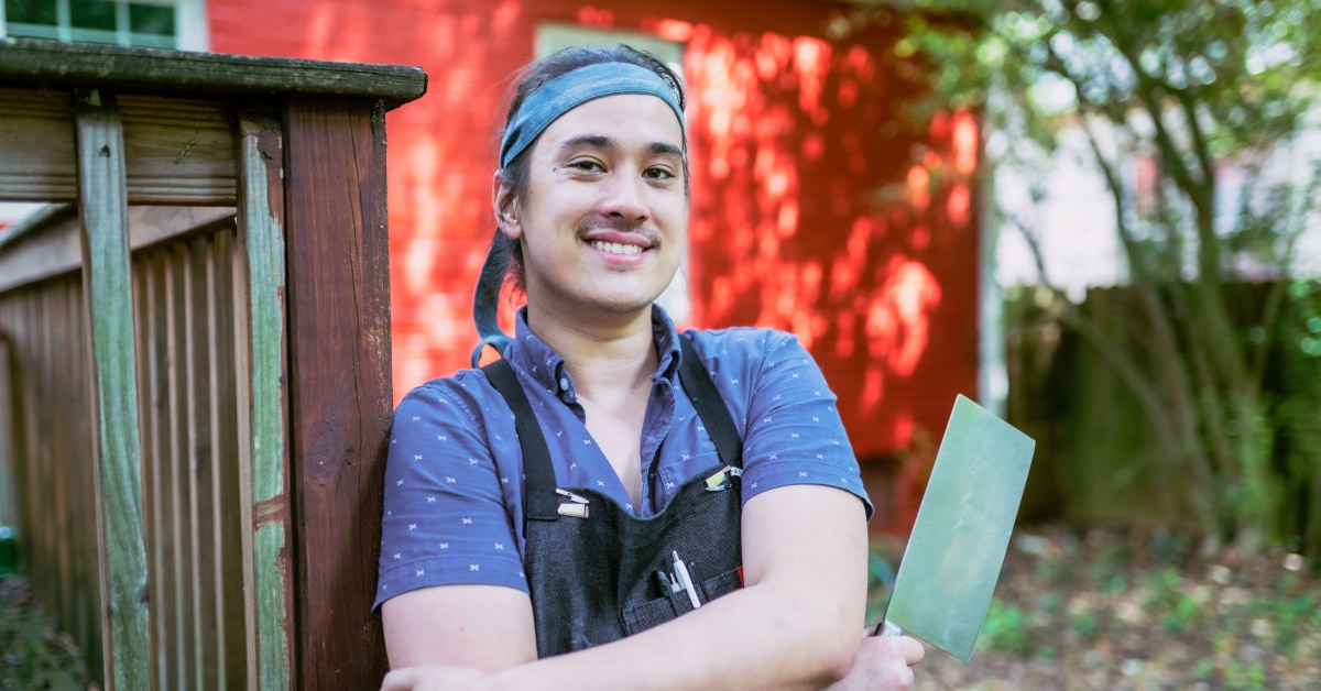 A young man stands in front of a red brick wall with a chef apron on and a sharpened cleaver.