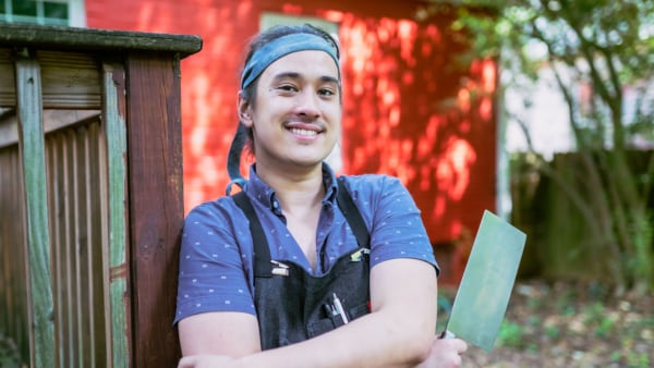 A young man stands in front of a red brick wall with a chef apron on and a sharpened cleaver.