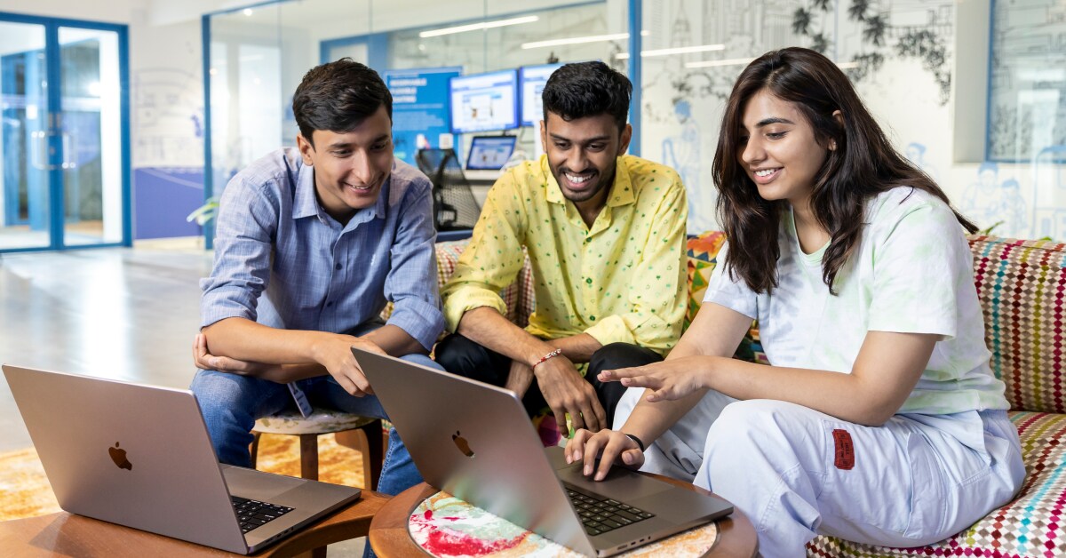 A group of Indian technology professionals collaborate around a laptop in a corporate office.