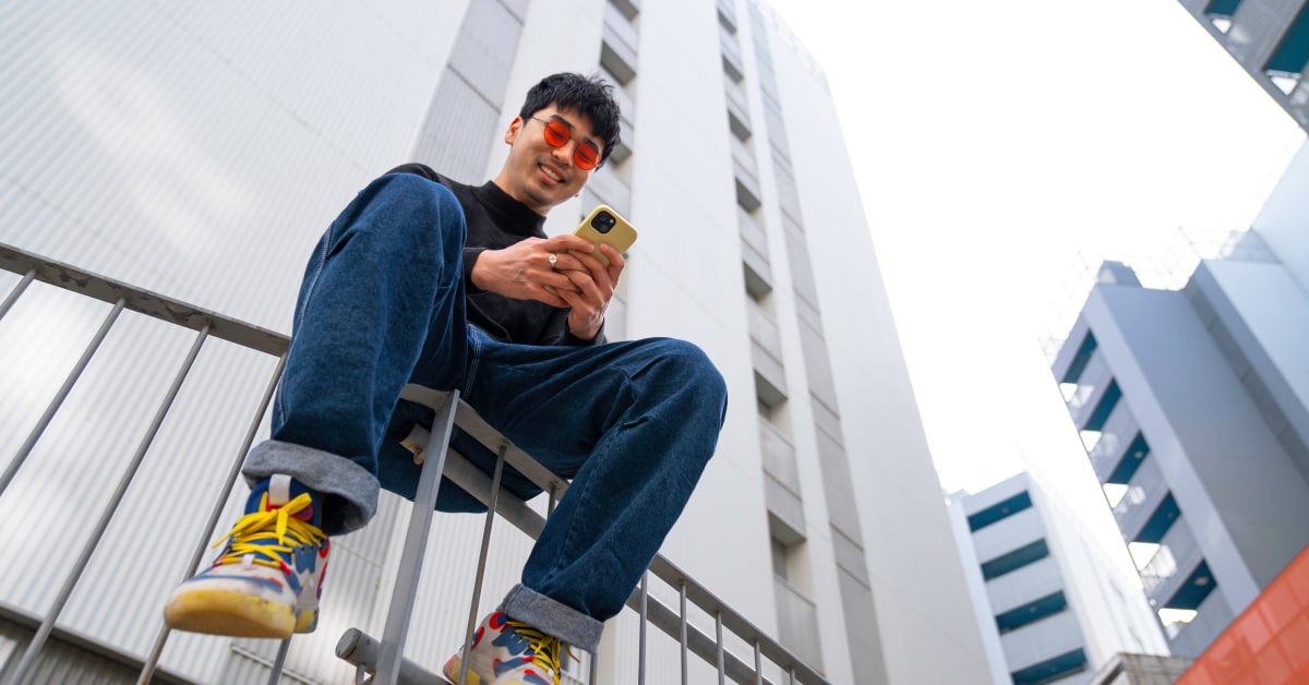 A young man sits on a fence in a city, looking at his phone.