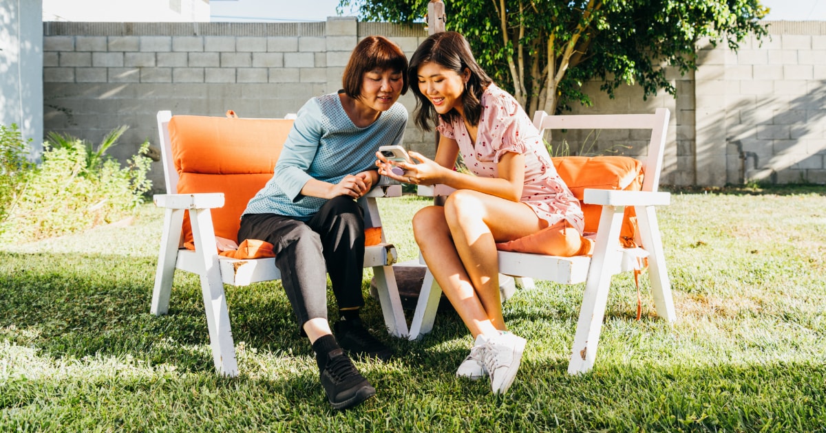 An Asian mother and daughter sit outside under an umbrella, looking at a phone and smiling.