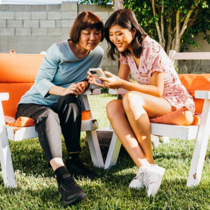 An Asian mother and daughter sit outside under an umbrella, looking at a phone and smiling.