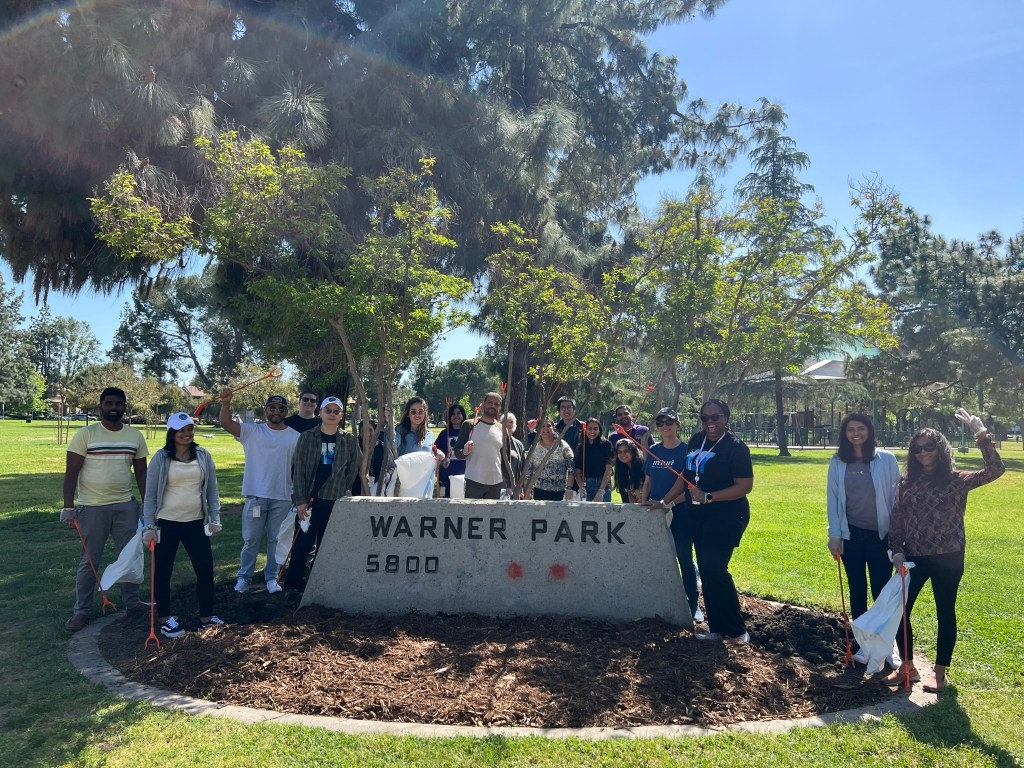 Volunteers stand around a stone park sign reading "Warner Park 5800" and smile with their garbage collecting tools.