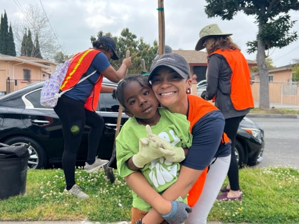 Volunteers in orange vests plant a tree in the background while a woman volunteer smiles with a Black child. 