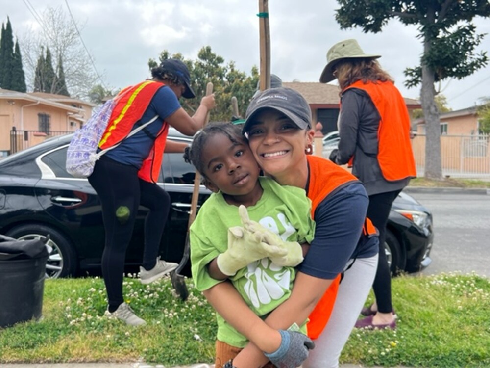 Volunteers in orange vests plant a tree in the background while a woman volunteer smiles with a Black child. 