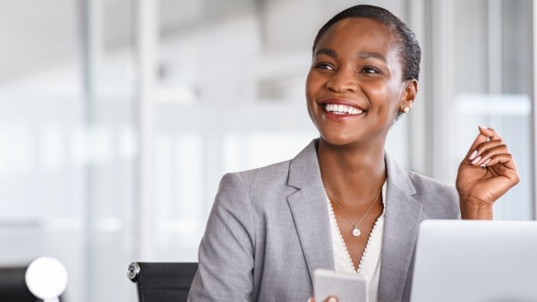 Smiling woman in a gray blazer sitting at a desk with a laptop and holding a smartphone in a bright, modern office