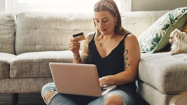 Woman sitting on a couch using a laptop and holding a credit card, with a small dog resting beside her
