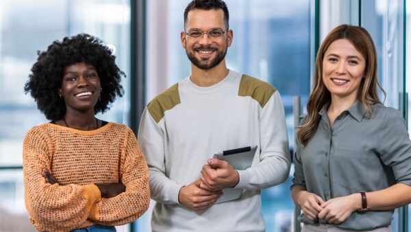 Three smiling coworkers standing together in a modern office; a woman in an orange sweater with arms crossed, a man holding a tablet, and a woman in a gray shirt