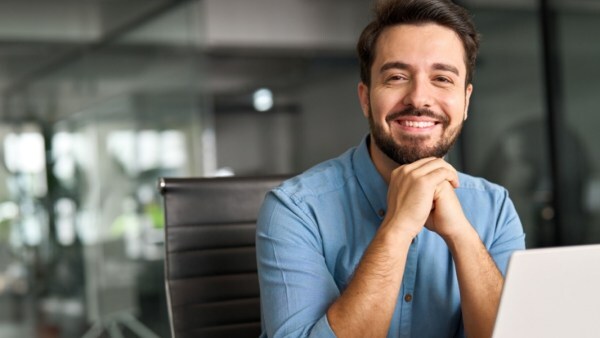 Smiling man in a blue shirt sitting at a desk with a laptop in a modern office.
