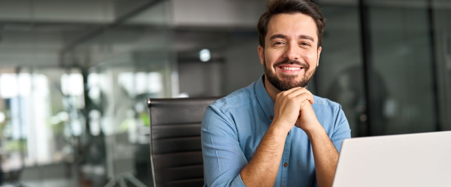 Smiling man in a blue shirt sitting at a desk with a laptop in a modern office.