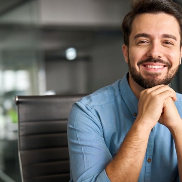 Smiling man in a blue shirt sitting at a desk with a laptop in a modern office.