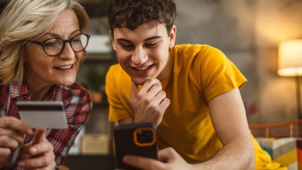 Smiling older woman holding a credit card while sitting with a teenage boy looking at a smartphone together in a cozy living room