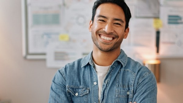 “Smiling man in a denim shirt standing with arms crossed in front of a wall covered with charts and notes