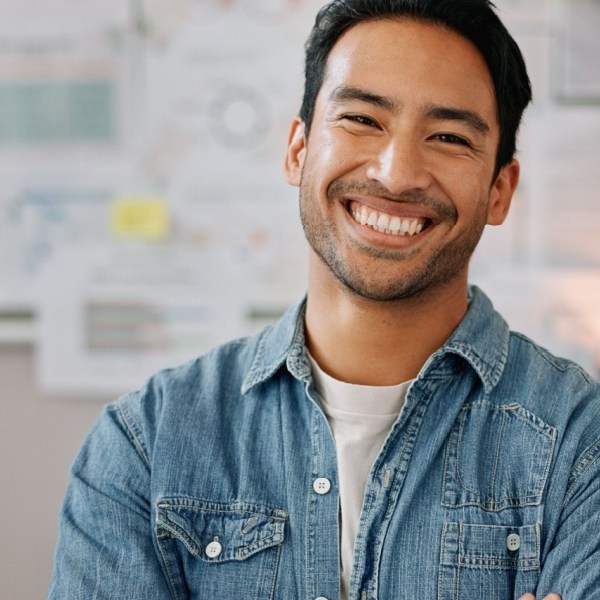 “Smiling man in a denim shirt standing with arms crossed in front of a wall covered with charts and notes