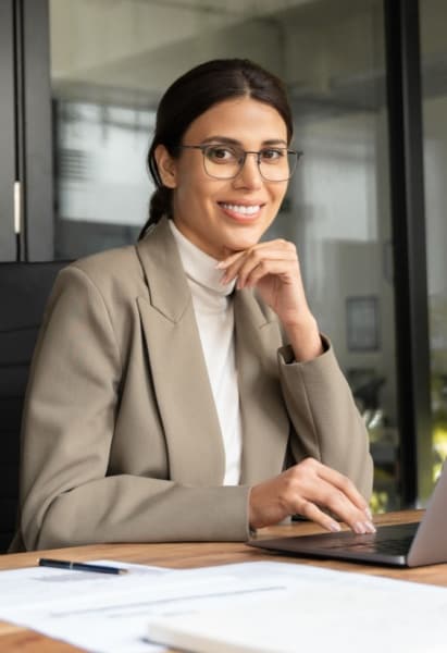 Smiling woman wearing glasses and a beige blazer sitting at a desk with a laptop in a modern office