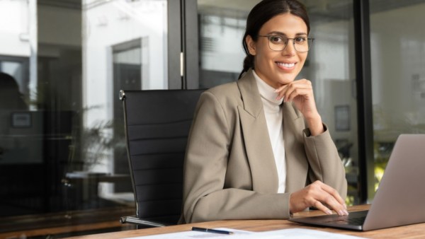 Smiling woman wearing glasses and a beige blazer sitting at a desk with a laptop in a modern office